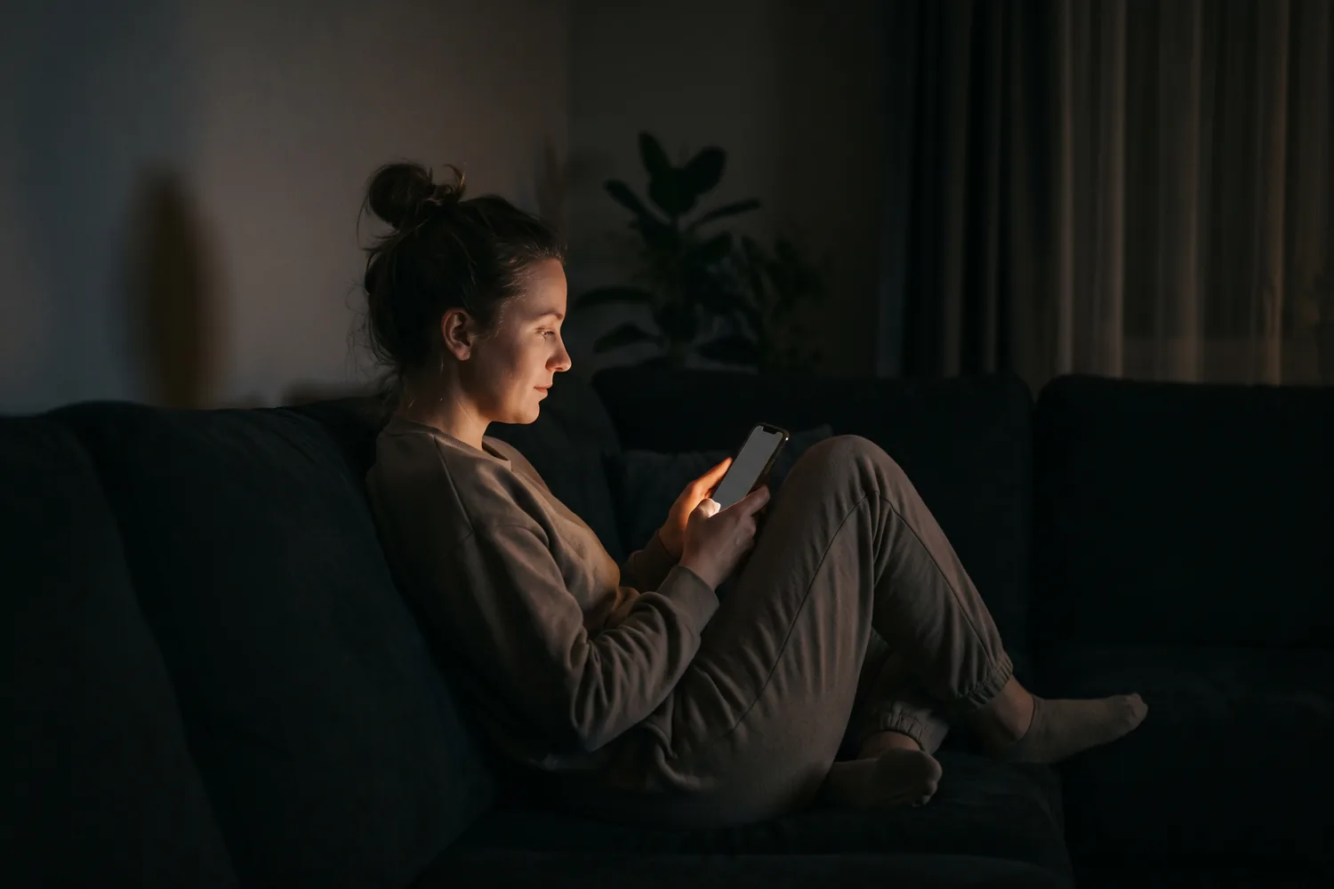 A woman sitting on a sofa in a dark living room at night, her face lit only by the phone she is holding.